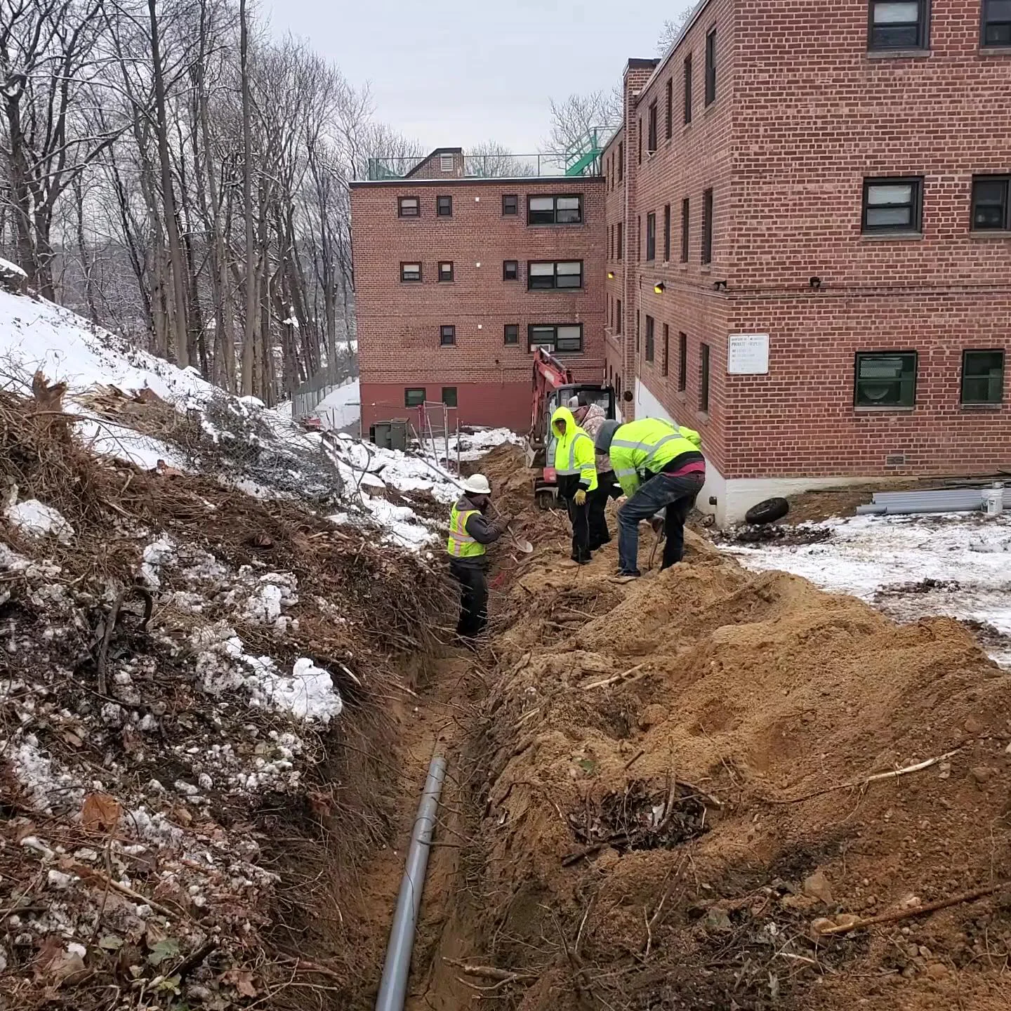 Four construction workers in safety vests and helmets are installing a pipe in a trench beside a brick building on a snowy day. Dirt is piled along the trench, and snow covers the surrounding ground.