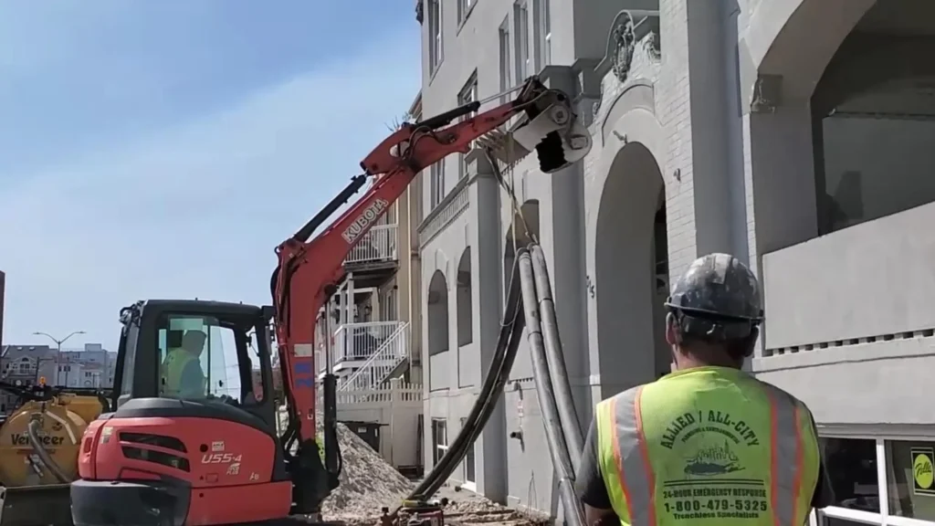 A construction worker in a safety vest and hard hat observes an excavator using its attachment to cut or break into the upper wall of a multi-story building under renovation or demolition.