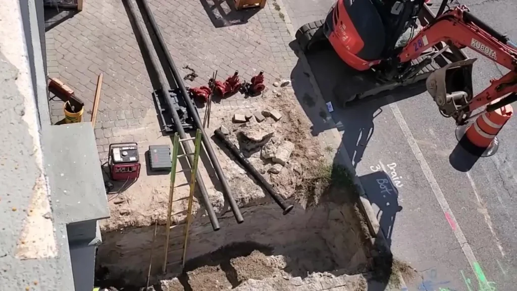 A construction site with a deep trench, pipes, a yellow ladder, scattered tools, and a red excavator. Orange traffic cones and roadside markings are visible near the excavation area.