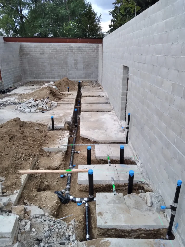 Construction site showing partially built concrete block walls and exposed plumbing pipes with blue caps coming out of the ground. Trenches run along the dirt and concrete floor, indicating ongoing plumbing installation.