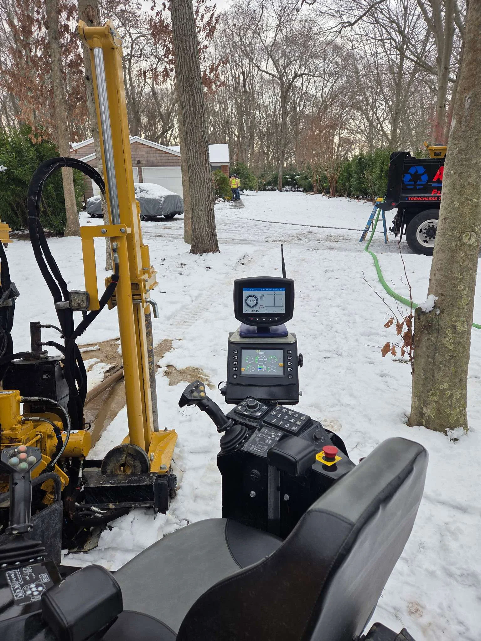 A control panel on a yellow drilling machine faces snowy ground in a wooded area. A black truck and cars are visible in the background, with bare trees and a house partially seen beyond the snow.