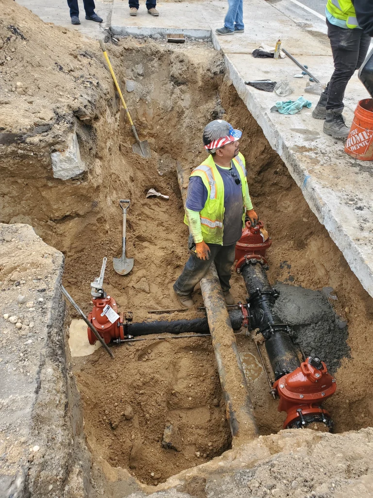 A construction worker in a safety vest and helmet stands in a dirt trench, surrounded by pipes and red valves, with shovels and tools nearby, working on underground utility maintenance.