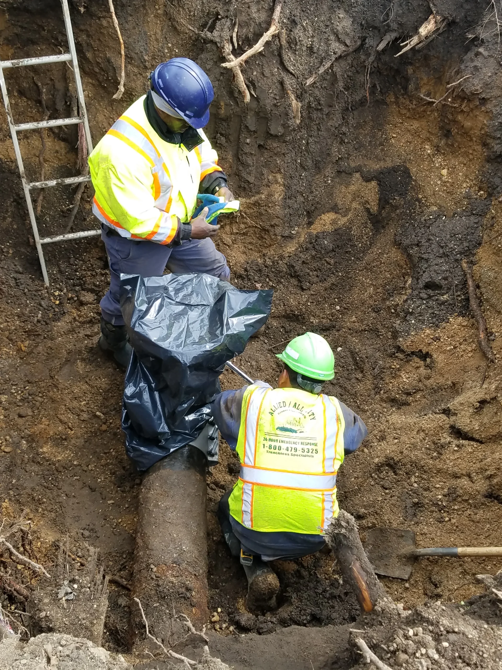 Two construction workers in safety gear and reflective jackets are working in a dirt trench. One stands and holds a tool and plastic sheet, while the other kneels beside a large pipe. A metal ladder leans against the trench wall.
