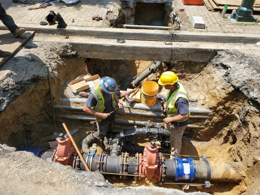 Two construction workers wearing safety gear and vests work in a large trench, installing or repairing underground pipes and valves. Tools, equipment, and pipes are visible around them inside the excavation site.