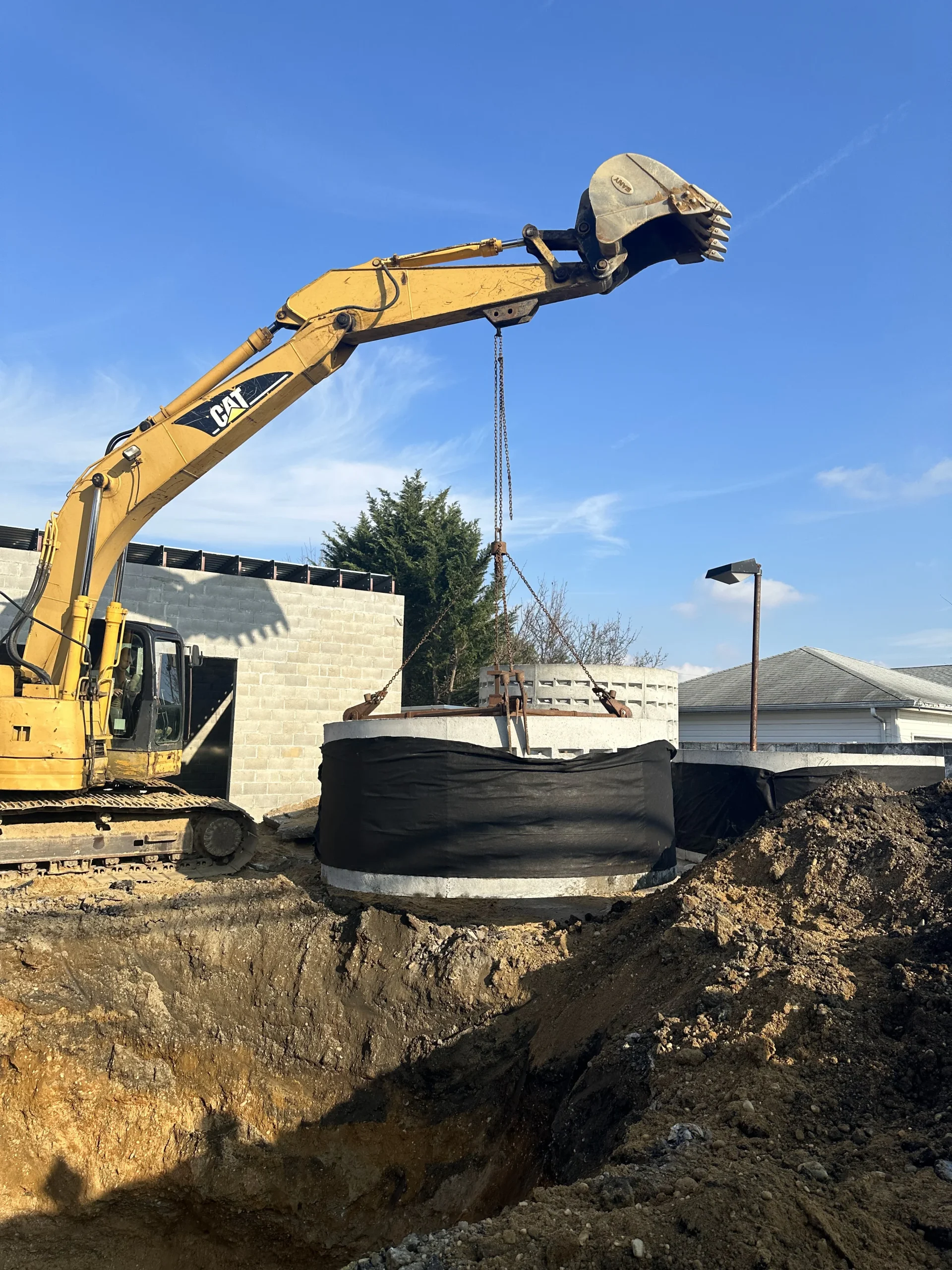 A yellow excavator lifts a large concrete cylinder wrapped in black plastic at a construction site, with a building and piles of dirt visible in the background under a clear blue sky.