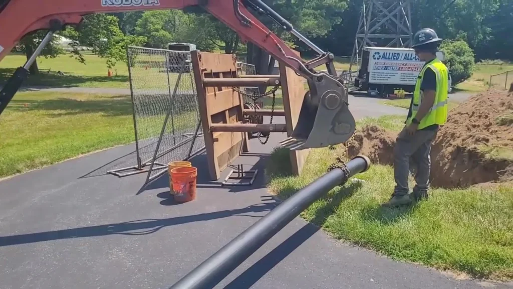An excavator lifts a large black pipe next to a dug trench on a paved road. A worker stands nearby, and a company truck and fencing are visible in the background on a sunny day.