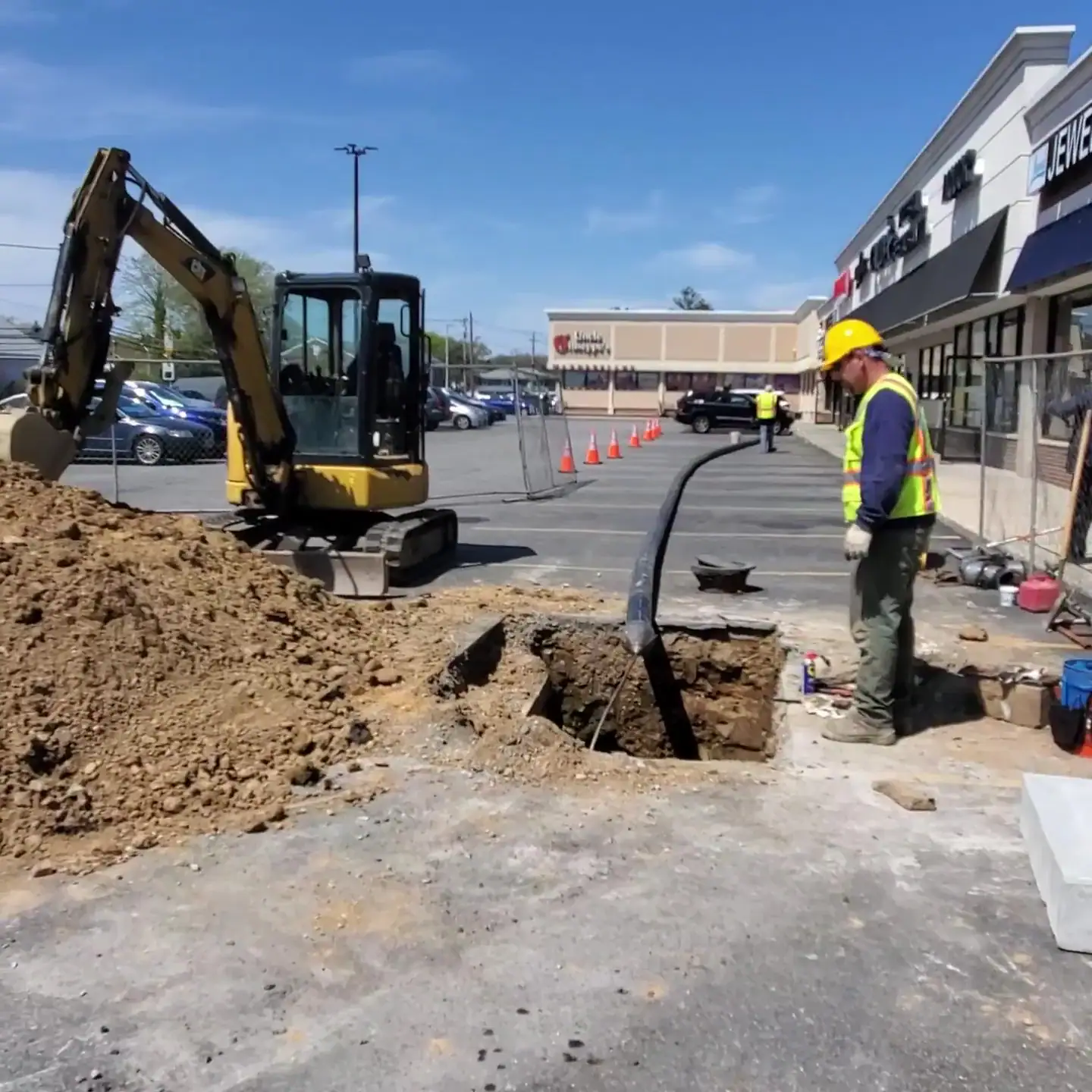 Workers in safety vests and helmets are operating near a hole in a shopping center parking lot, with a small excavator and a large dirt pile nearby. Stores and traffic cones are visible in the background.