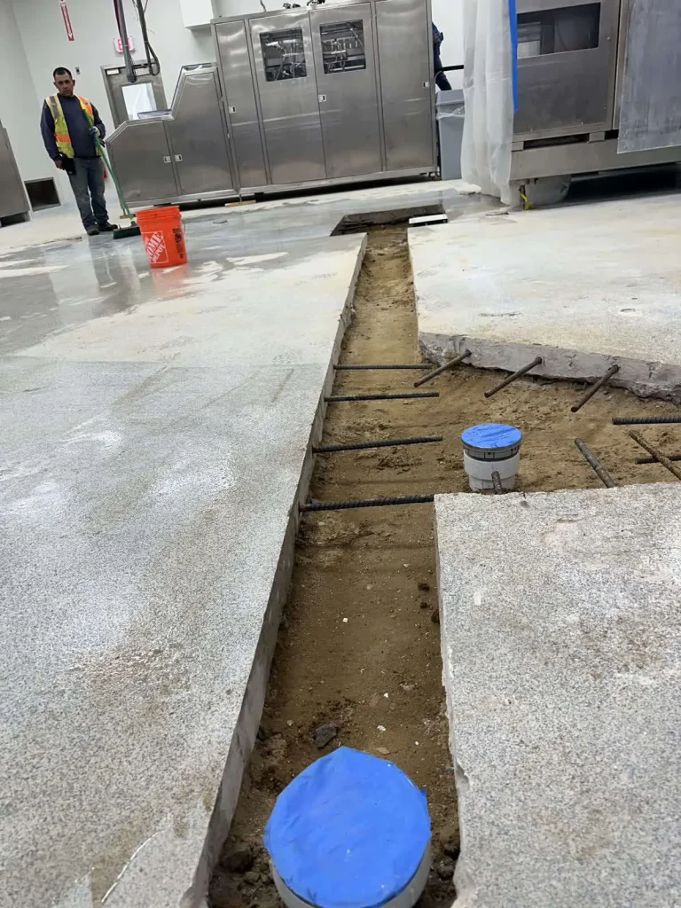 A construction site indoors shows a trench cut into the concrete floor with exposed dirt and rebar, plastic-covered pipes, and large machinery in the background. A worker and an orange bucket are visible on the left side.