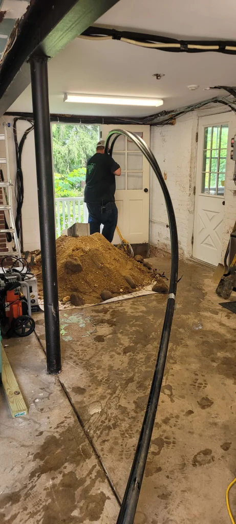 A person stands in a partially dug-up room next to a pile of dirt, near double doors leading outside. Construction tools and materials are scattered around, and a large black cable arches through the space.
