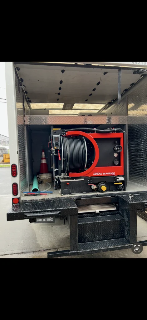 A truck with its rear door open reveals equipment inside, including a large red and black hose reel labeled "Urban Warrior," orange traffic cones, and additional tools stored beside it.