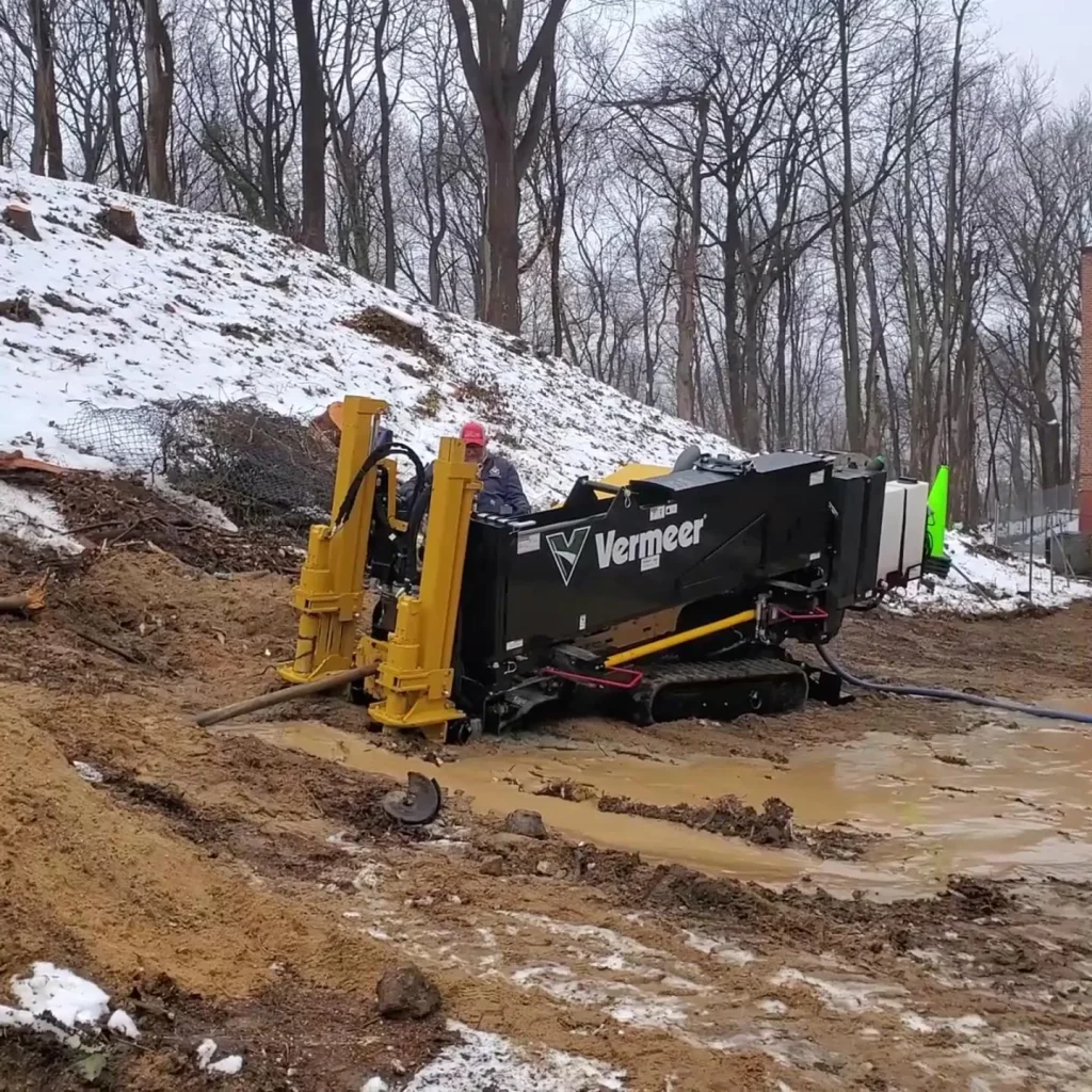 A Vermeer horizontal directional drill sits on muddy ground at a construction site near a snowy hillside, surrounded by leafless trees in winter.