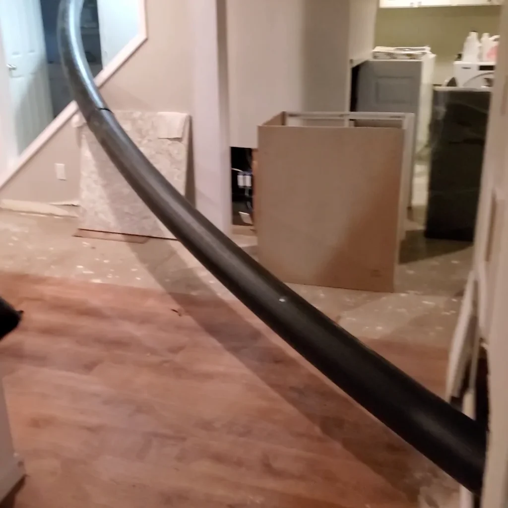 A long, black pipe or tube runs diagonally through a house under renovation, stretching from an upper level to the ground floor with bare subfloor and construction materials visible.