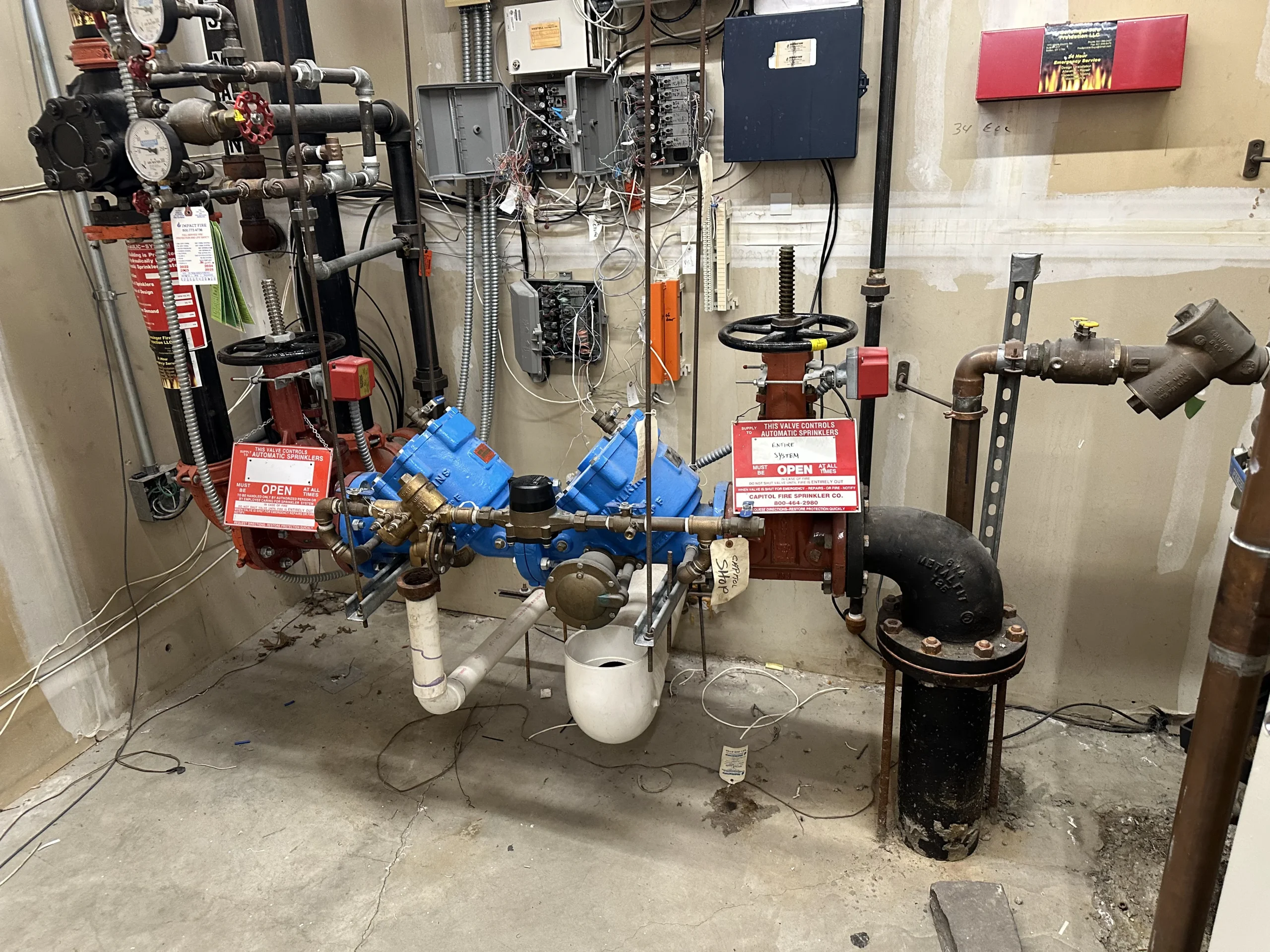 A basement utility room with exposed pipes, gauges, and valves, including two red-handled valves labeled "OPEN." Electrical panels and wiring are mounted on the back wall, and a red emergency alarm box is visible.