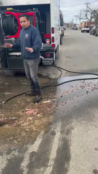 A person in a dark sweatshirt and jeans stands on a wet, muddy street near a work truck with hoses, raising their hands slightly. Water and debris are visible on the ground.