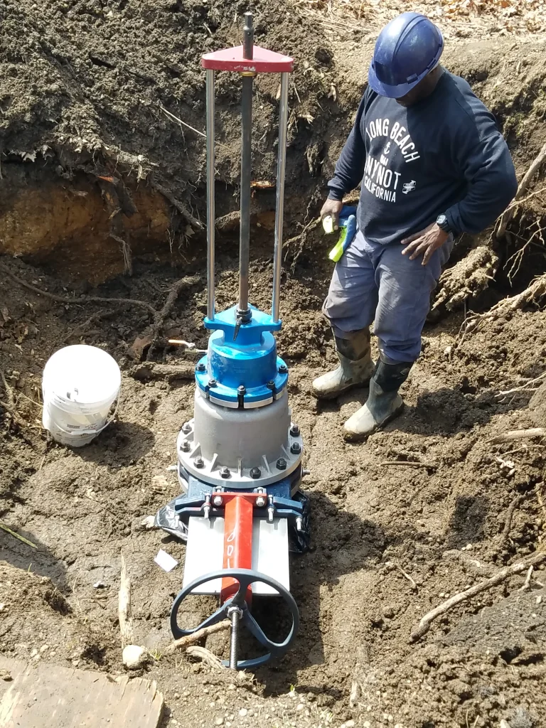 A worker wearing a helmet and boots stands next to a large underground valve installation in a dirt trench, holding a tool and inspecting the equipment.