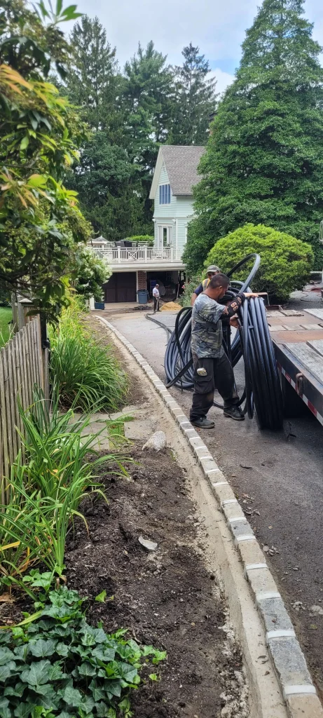 A person in work clothes unloads coiled black tubing from a flatbed truck on a paved driveway, with a house, trees, and a sloped garden bed nearby. Another person stands further up the driveway.