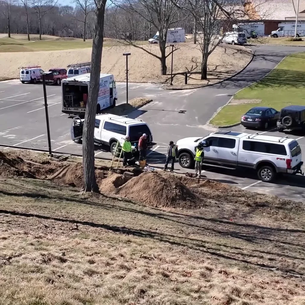 Several utility workers wearing safety vests dig trenches along a grassy hill near parked utility vehicles in a parking lot. The area is surrounded by dry grass and a few trees, with buildings in the background.