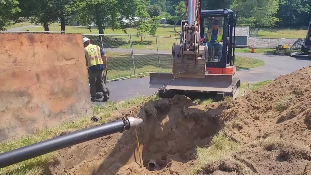 Two construction workers operate excavation equipment near a trench, where a black pipeline is being installed underground. Dirt piles surround the trench, and a fence encloses a grassy park area in the background.