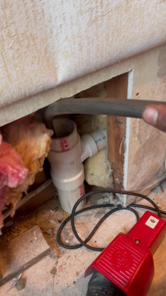 A hand holds a black hose near exposed PVC pipes and insulation inside a wall opening, with a red tool and black cable on the floor below.