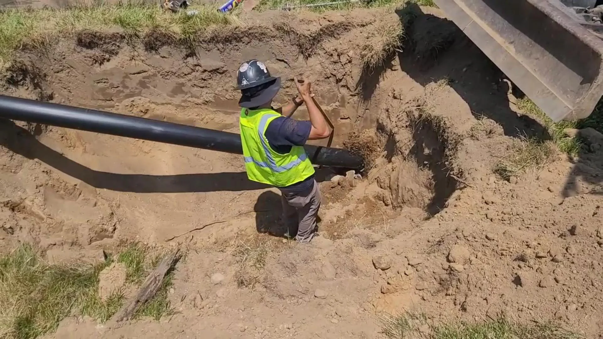 A construction worker in a safety vest and helmet stands in a sandy trench, guiding a large black pipe into place. Excavated soil surrounds the trench, and construction equipment is partially visible at the edge.
