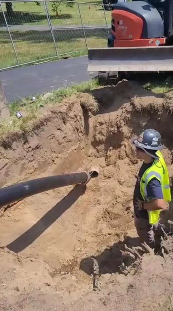 A construction worker in a safety vest and helmet stands in a large sandy pit, next to two exposed pipes. Excavator tracks and a metal fence are visible in the background.