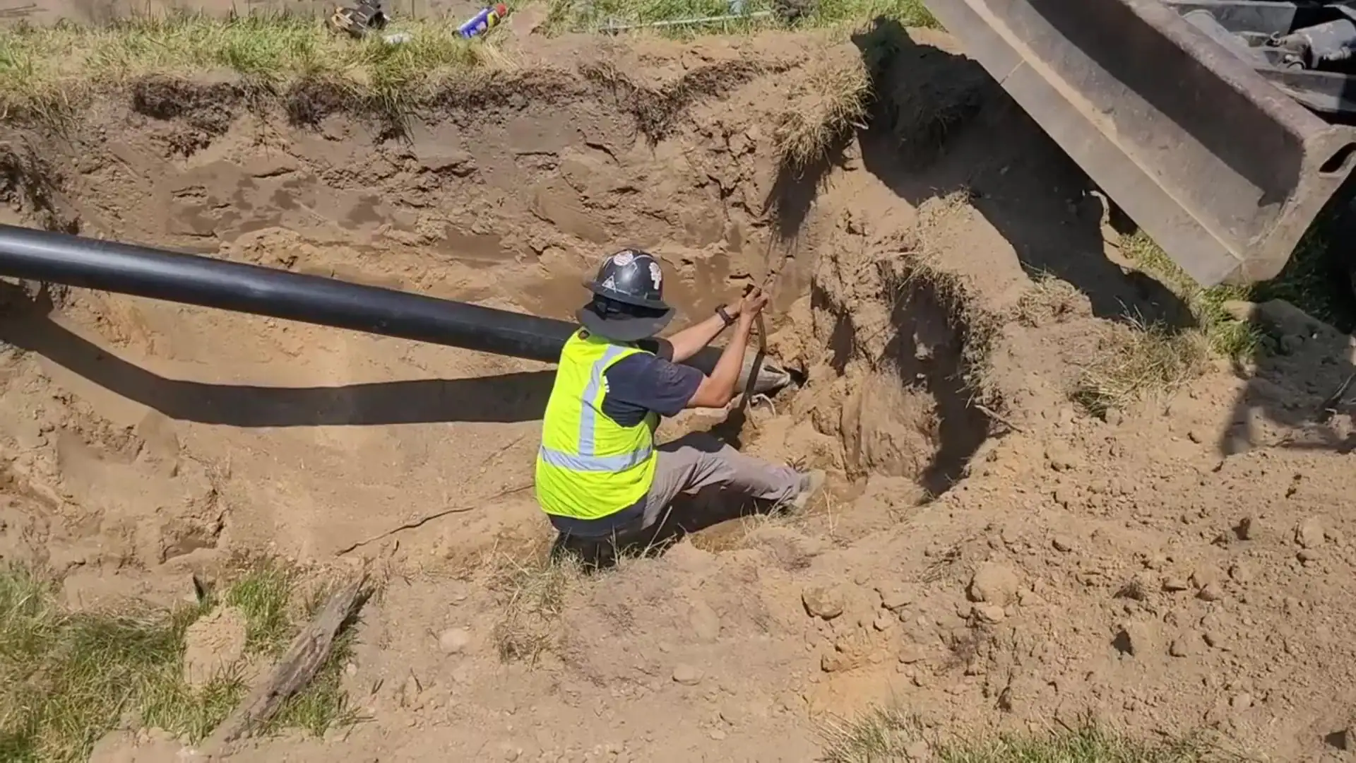 A construction worker in a yellow safety vest and helmet kneels in a trench, positioning a large black pipe while surrounded by dirt and construction equipment.