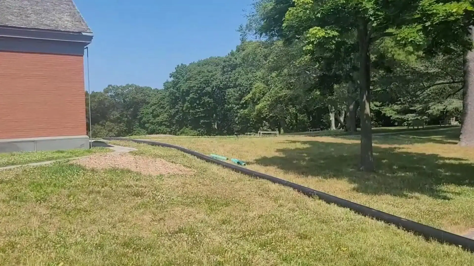 A grassy field with a black pipe or hose running across it, bordered by trees and a building on the left under a clear blue sky.