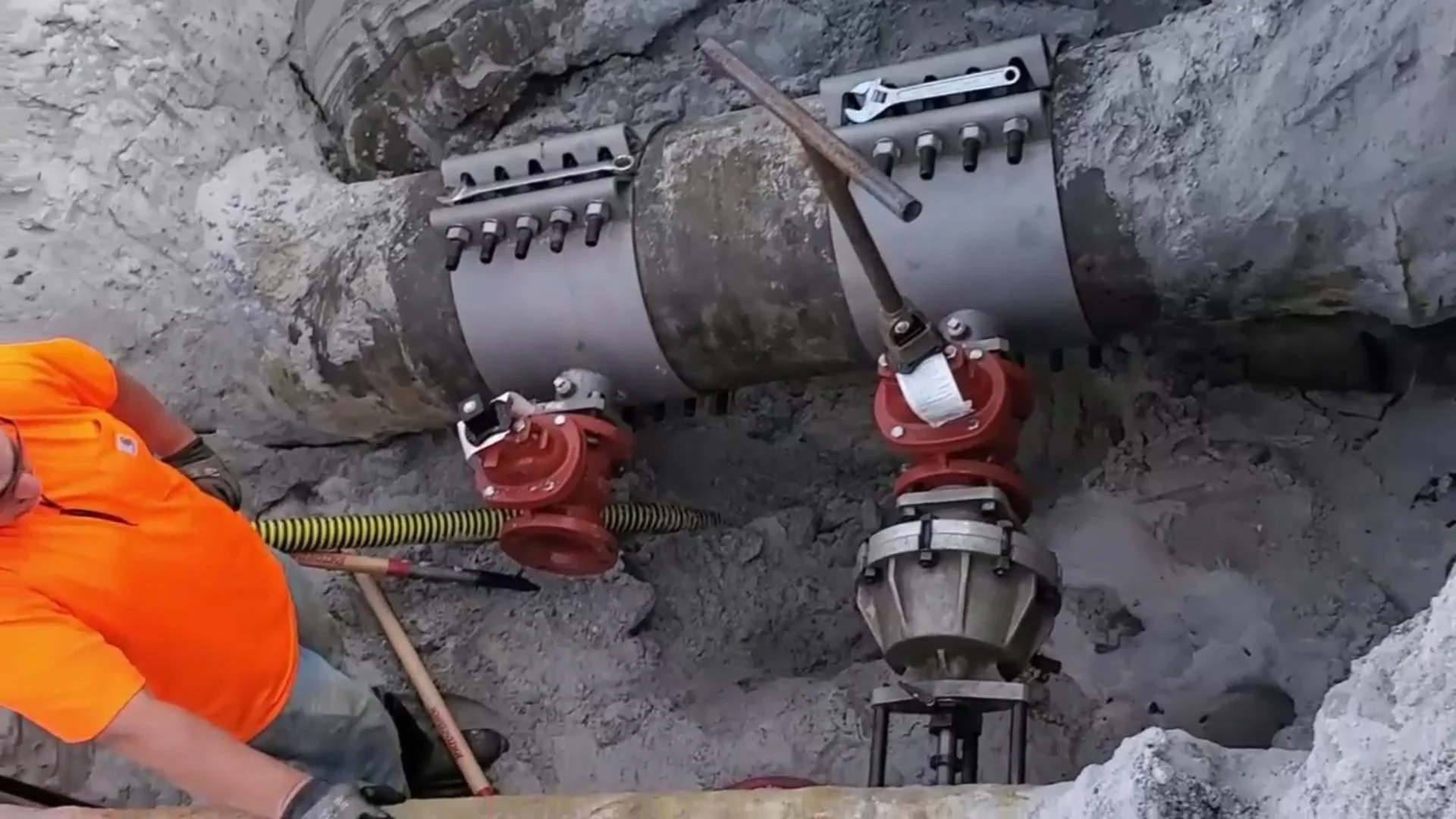 Two large metal pipe clamps secure a section of underground pipe. Two red valves and a large mechanical tool are attached to the pipe. A worker in an orange shirt is partially visible on the left side.