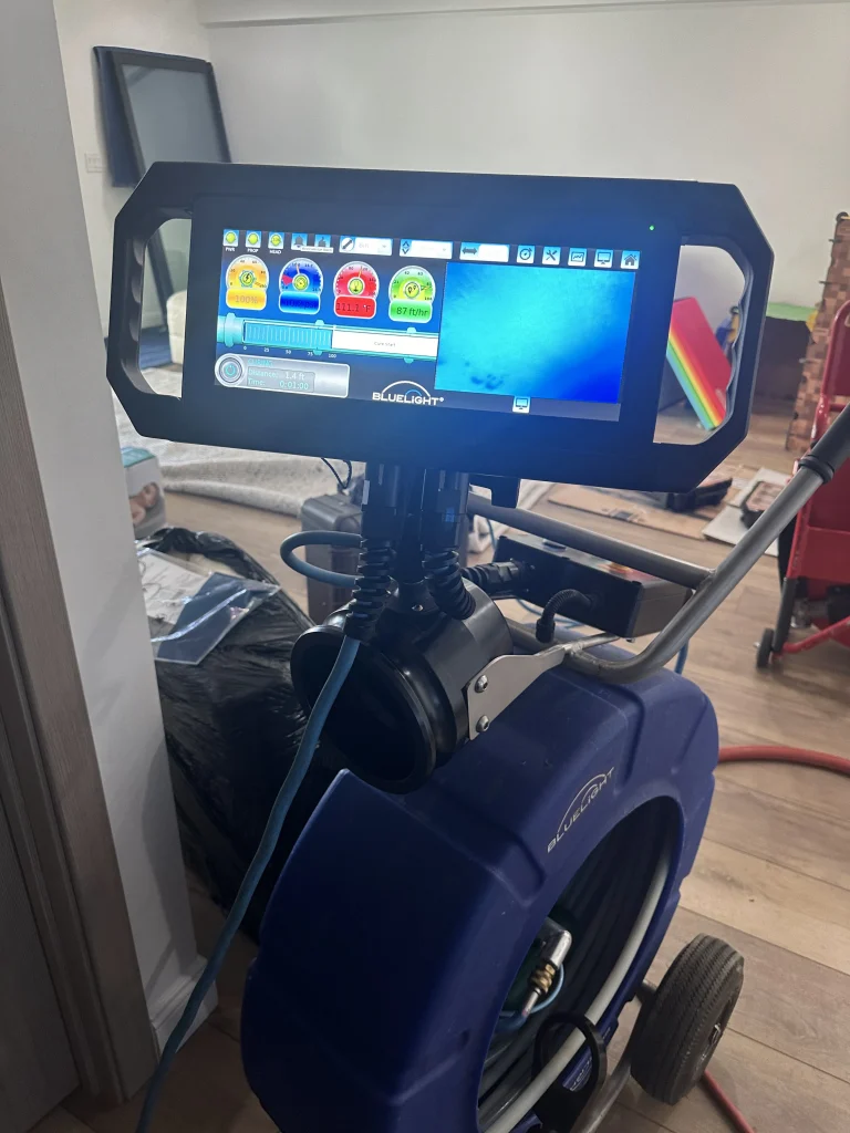 A close-up of a large blue industrial machine with a touchscreen control display, showing colorful icons and settings. The machine is on wheels and is set up indoors on a wooden floor.