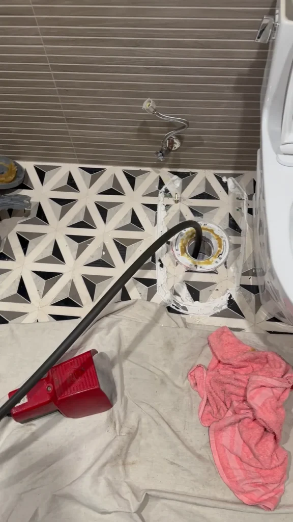 A bathroom with a toilet removed, exposing the toilet flange. A black hose or pipe is inserted into the flange. There is a red dustpan, a rag, and tools on a white sheet covering the patterned tile floor.