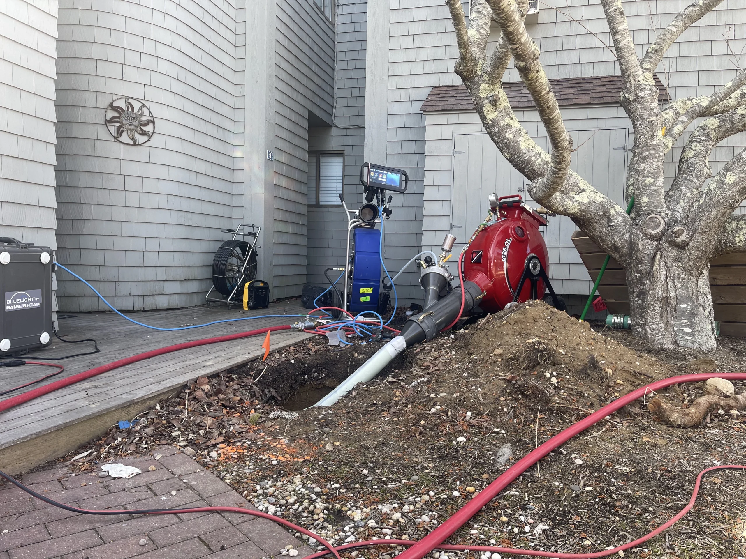 A large red vacuum excavation machine and hoses are set up outside a gray wooden building next to a tree. Soil has been dug up, exposing pipes, and equipment is spread across the yard and walkway.