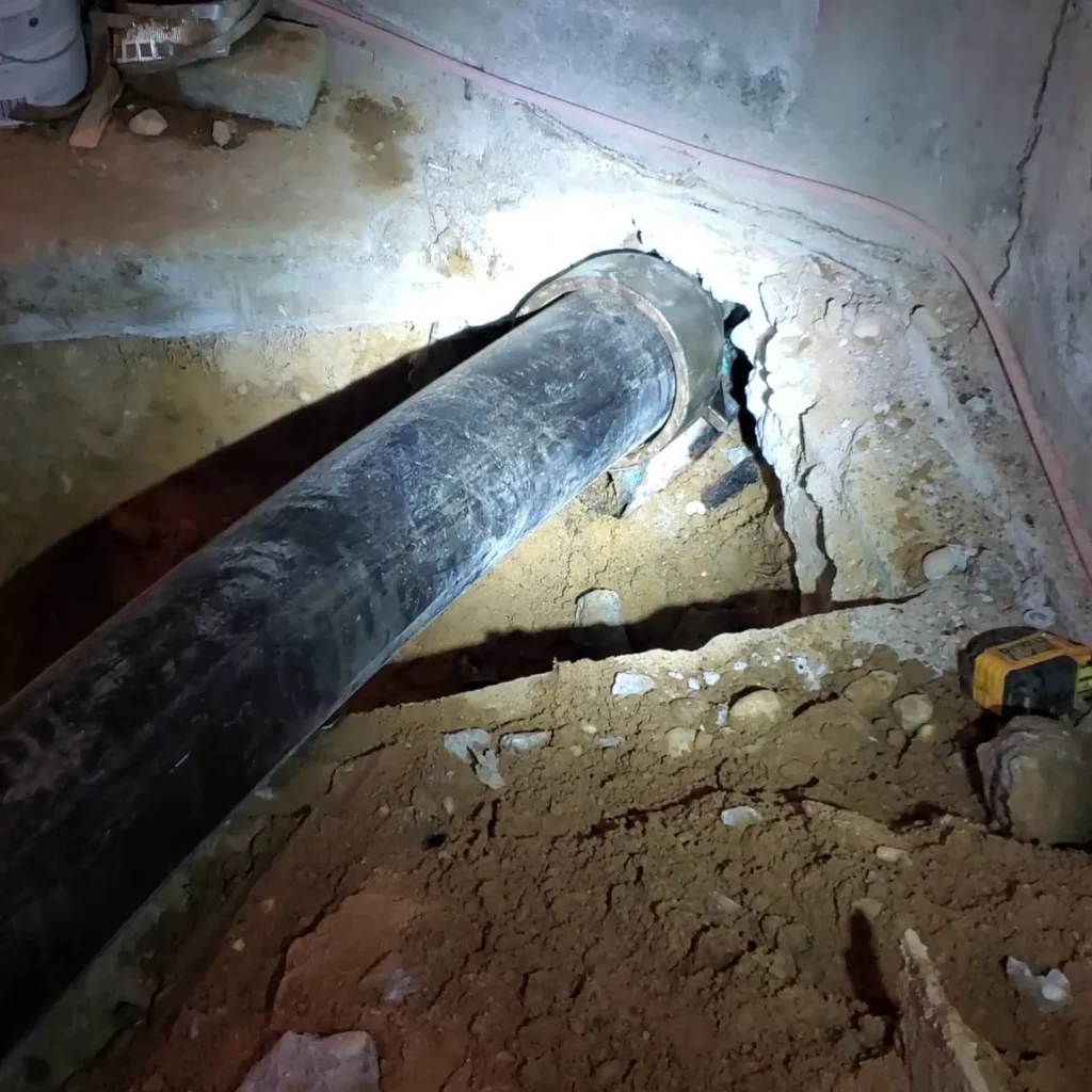 A large black pipe runs through a dirt floor, entering a wall with cracked concrete around the opening; a flashlight illuminates the area and a yellow measuring tape lies nearby.