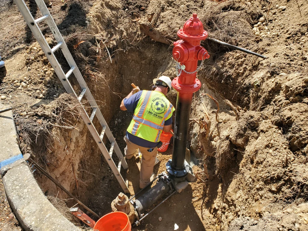 A worker wearing a safety vest and hard hat installs or maintains a red fire hydrant in a dug-out trench, with a ladder positioned nearby and construction tools and materials around.