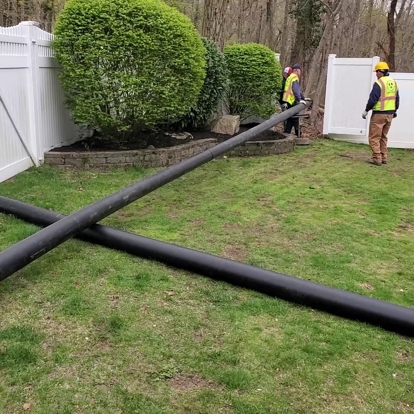 Two workers in safety vests and helmets stand near two large black pipes lying on a grassy yard beside a white fence and trimmed bushes.