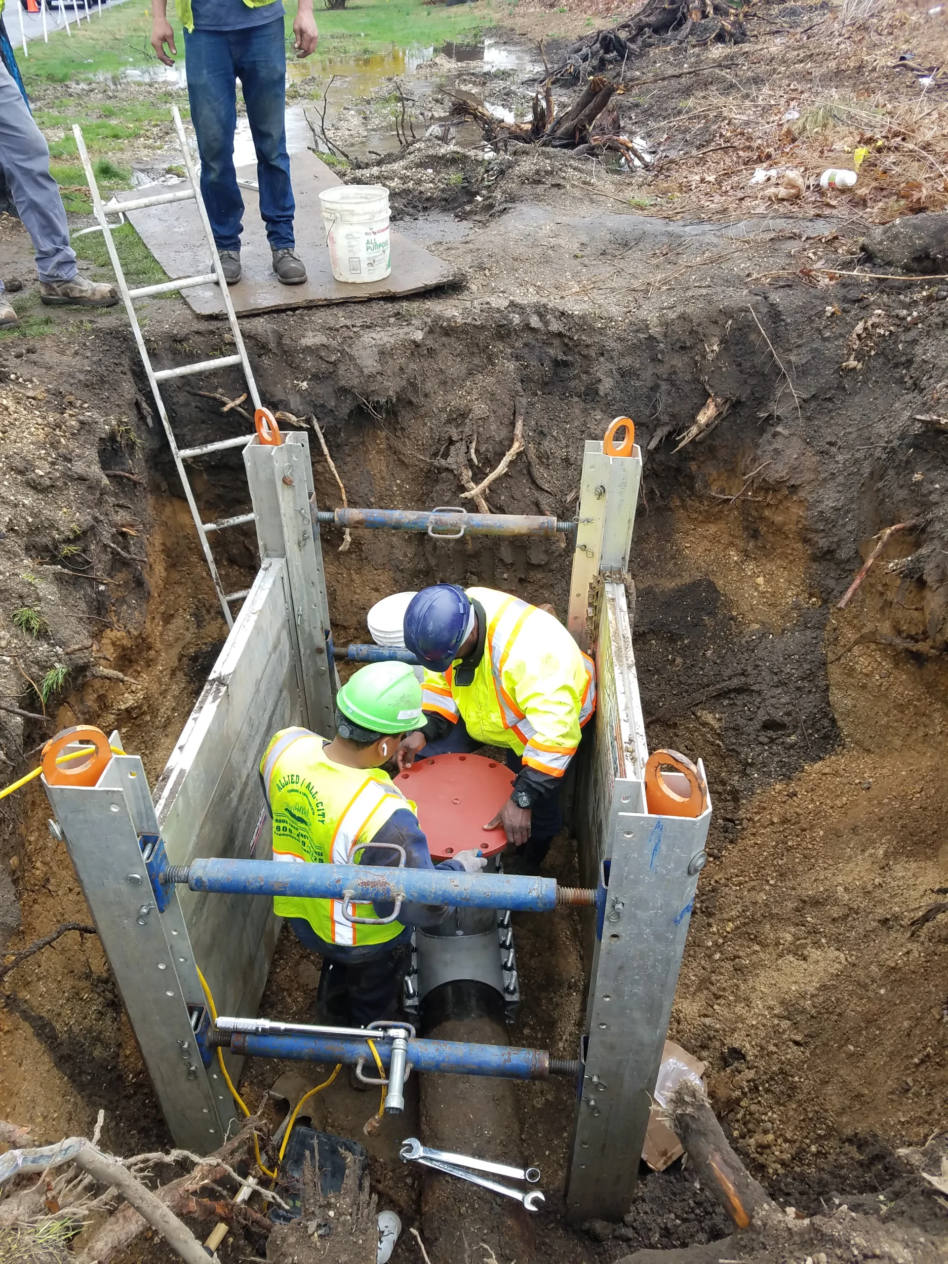 Two construction workers in safety gear are inside a trench box, installing a red valve onto a large black pipe underground, while two other workers stand above ground near a ladder and a bucket.