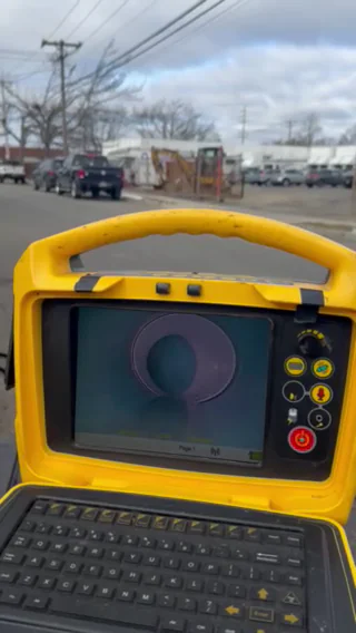 A close-up of a yellow electronic device with a keyboard and screen displaying a purple omega (Ω) symbol, set outdoors near a street with cars and utility poles in the background.