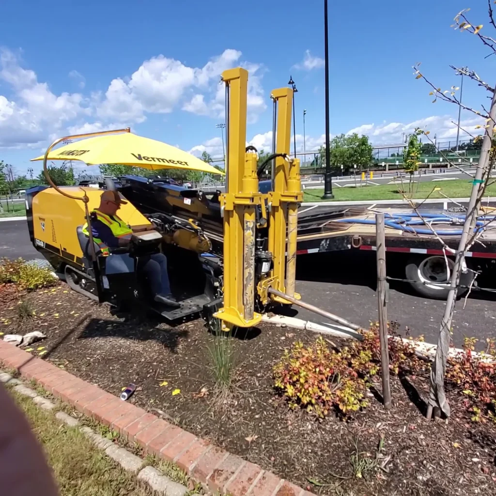 A worker operates a yellow Vermeer horizontal directional drilling machine on a landscaped area next to a brick border, with a truck and trees in the background under a blue sky with clouds.