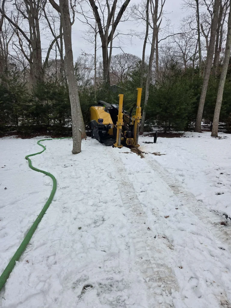 A yellow drilling machine is parked on snowy ground near a line of green trees. A long green hose runs along the snow, leading toward the equipment. Tire tracks are visible in the snow.