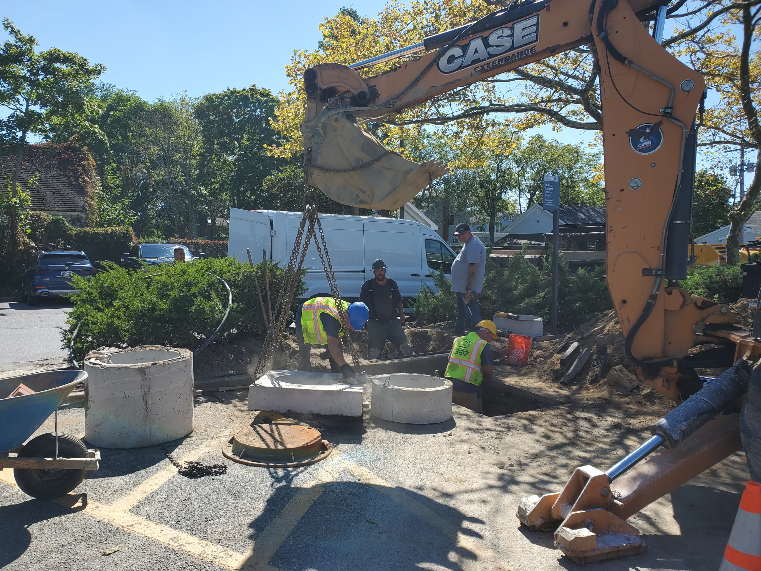 Construction workers wearing safety vests and helmets use an excavator and chains to lift concrete rings at a worksite in a parking lot. A white van and orange cone are visible in the background.
