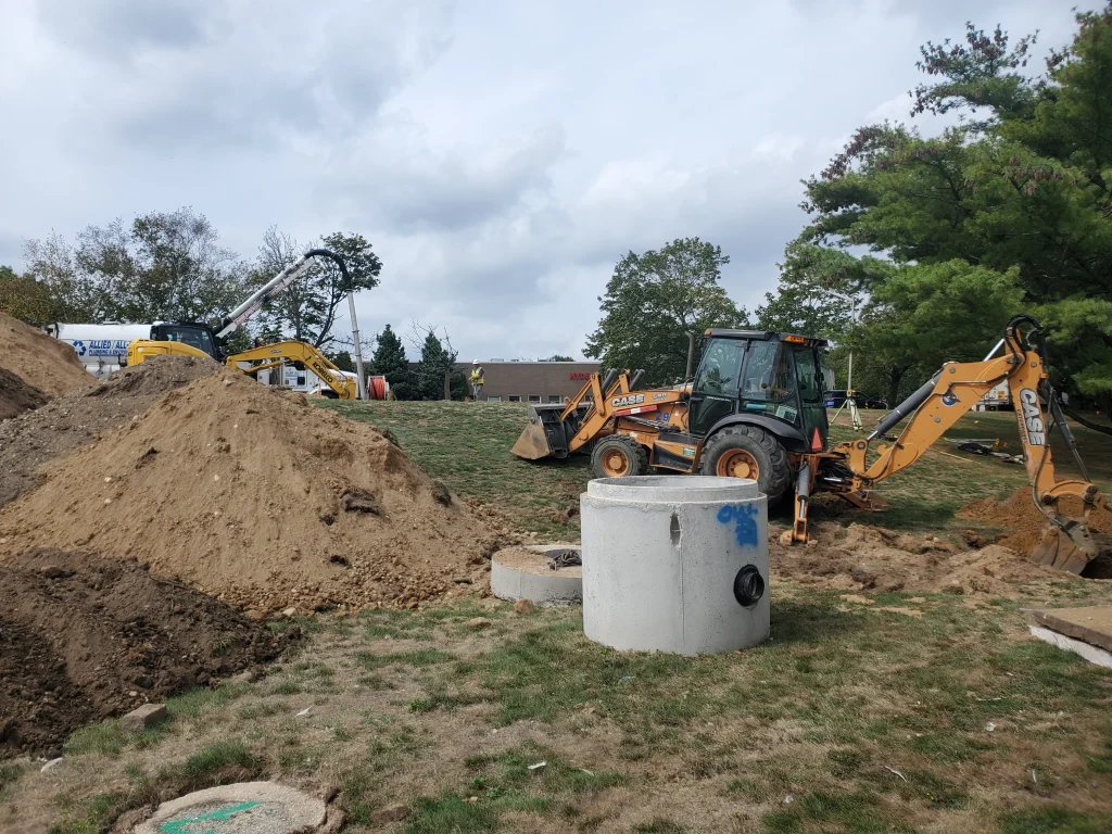A construction site with piles of dirt, a concrete cylinder structure, two excavators, and trees in the background under a cloudy sky.