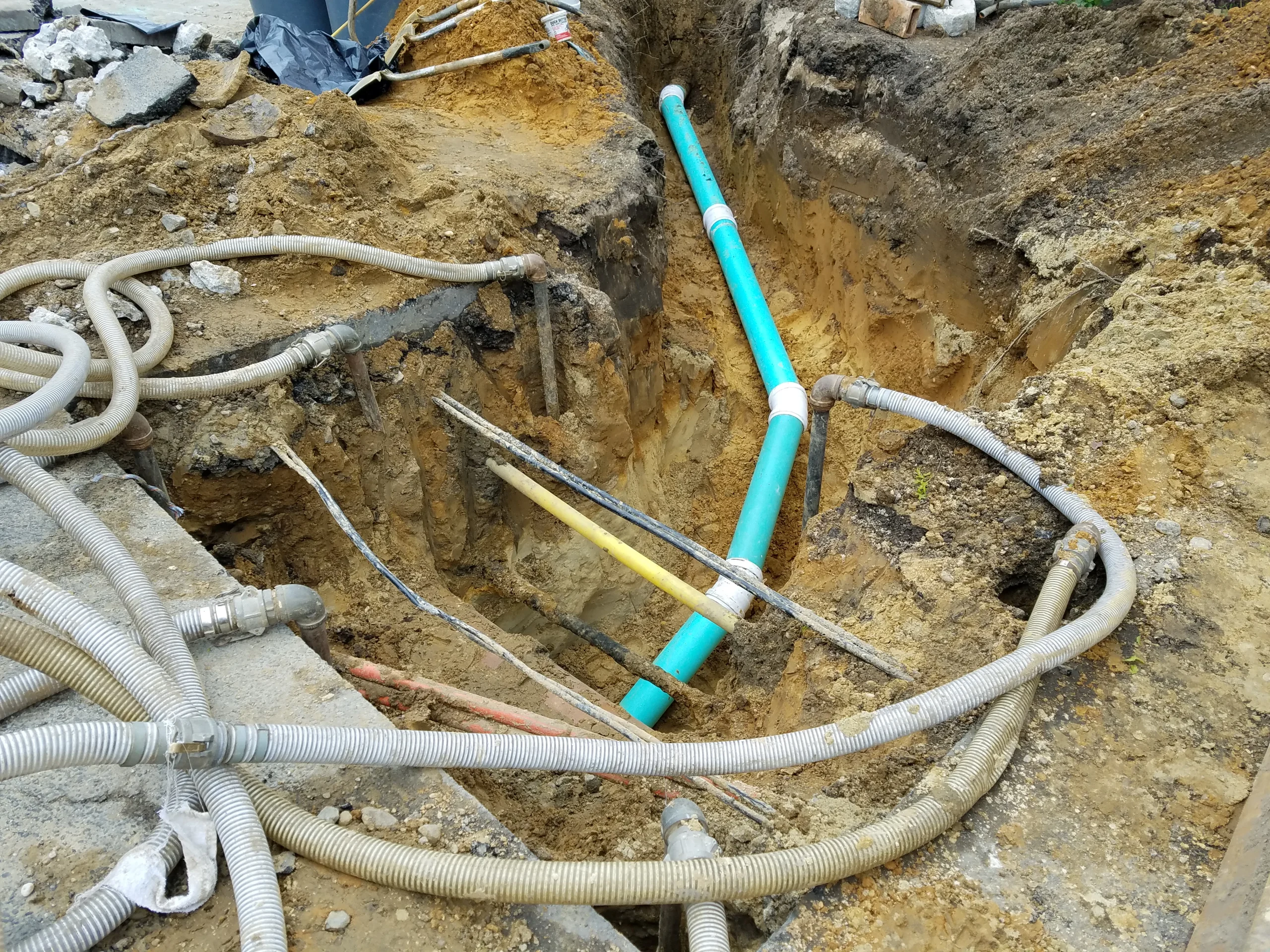 A construction site trench with exposed blue and yellow utility pipes, surrounded by dirt and various cables running across and into the trench.