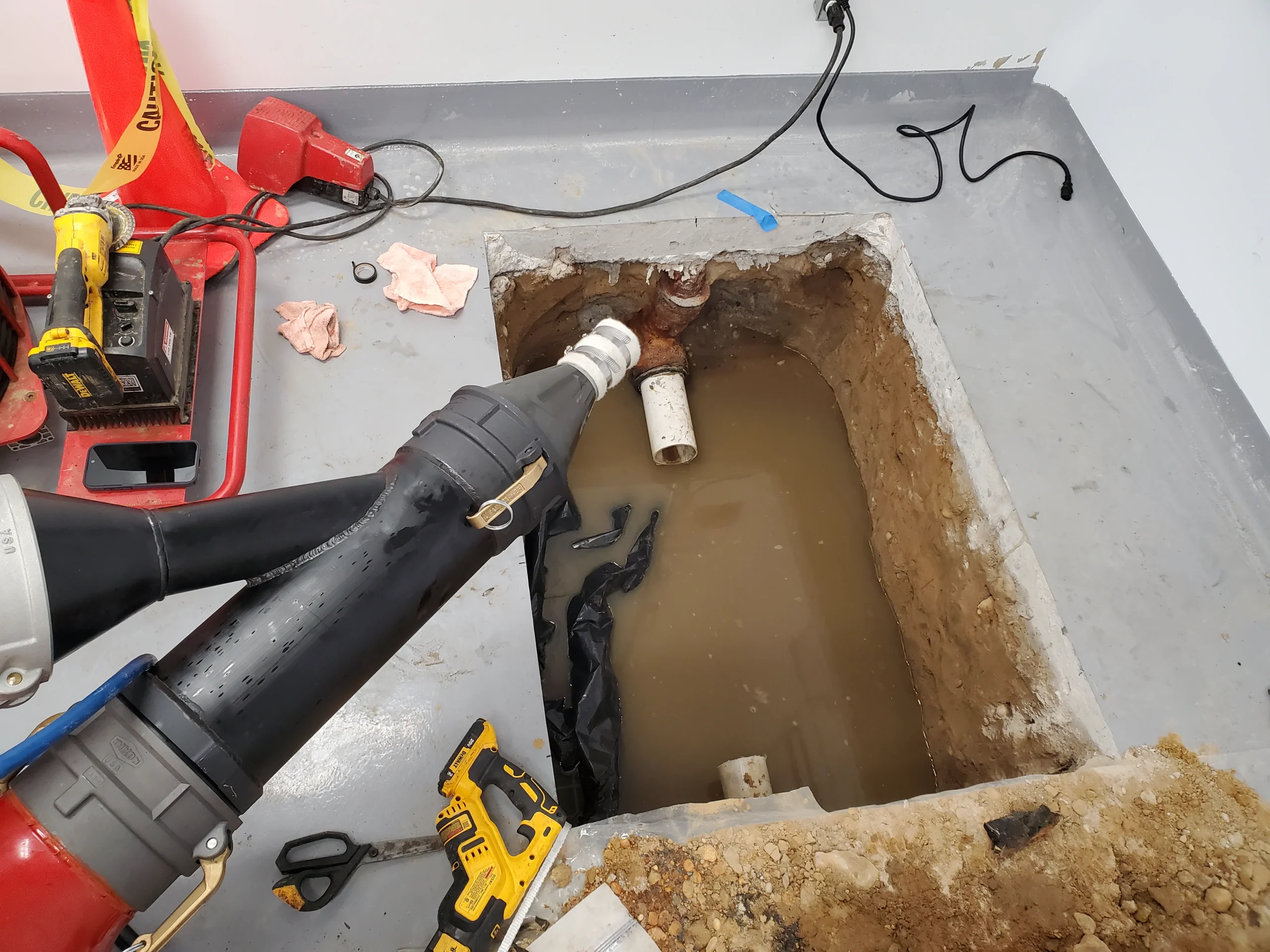 A large hole in a concrete floor reveals muddy water and exposed pipes. Plumbing tools, a drill, and other equipment are scattered nearby, indicating ongoing plumbing or repair work.