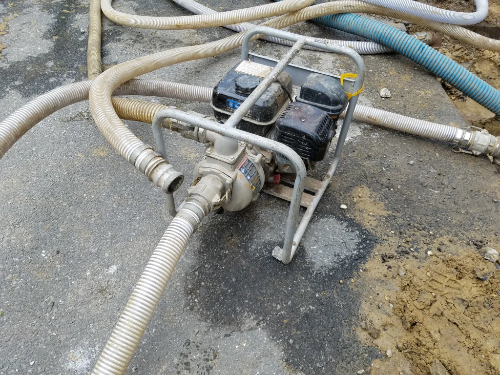 A portable water pump with attached hoses placed on a rough, partially muddy surface, surrounded by coiled hoses of various colors.