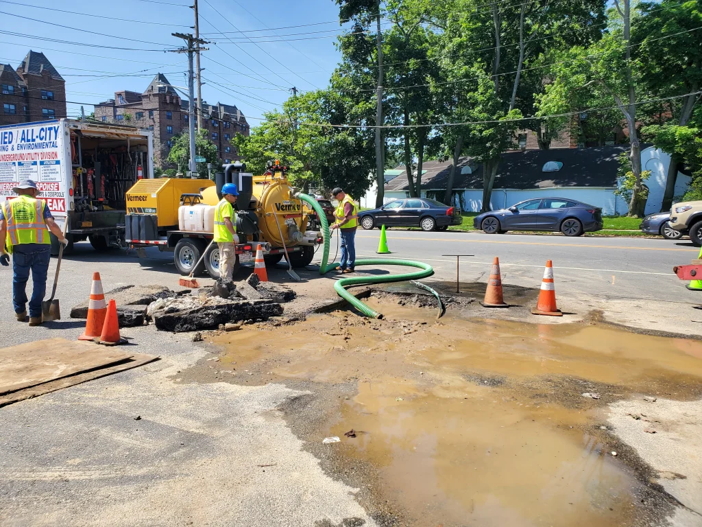 Workers in safety vests repair a water main break on a city street. Traffic cones, equipment, and a large puddle surround the work area. A yellow truck and several parked cars are in the background.