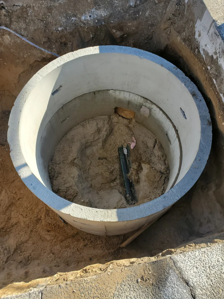 A concrete manhole ring is installed in the ground with sand and soil inside. Two partially buried cables or pipes are visible at the center. The surrounding area shows excavation work.