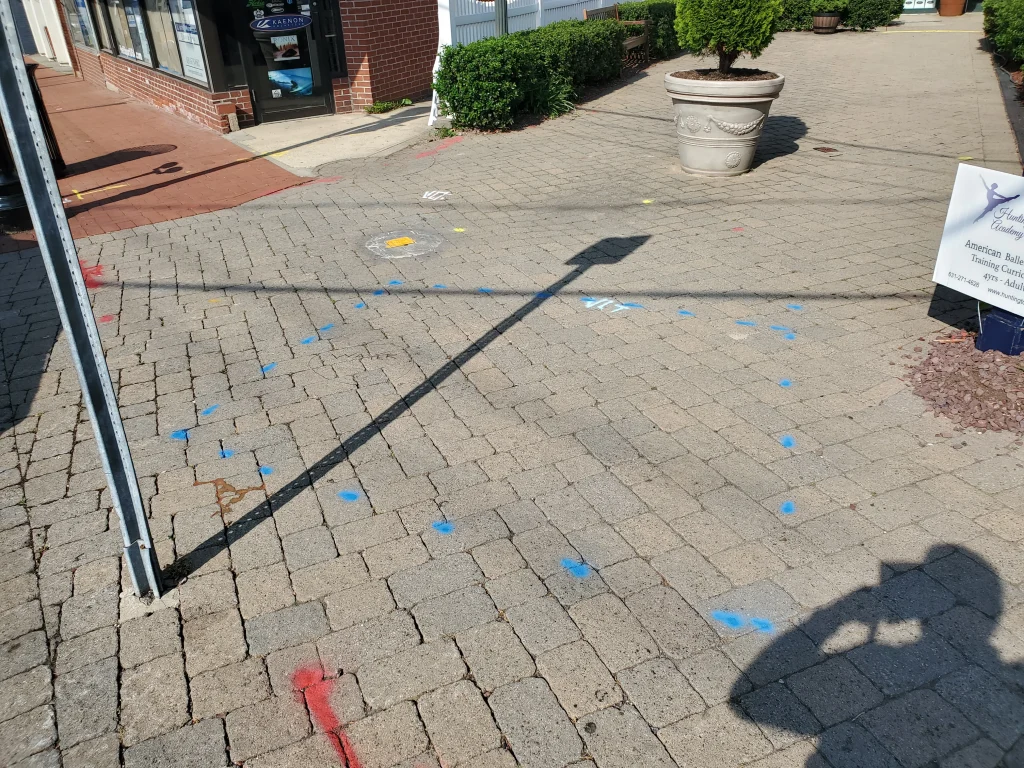 A brick sidewalk with scattered blue, red, and yellow utility markings. A large planter and building entrance are visible, along with a shadow of a street sign and a person taking the photo. A business sign rests nearby.