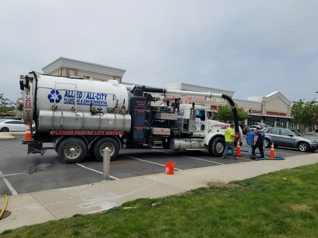 A large Allied All-City Plumbing & Environmental truck is parked in a lot near retail stores. Three workers in safety gear stand beside the truck, which has hoses attached. Orange cones and buckets are nearby.