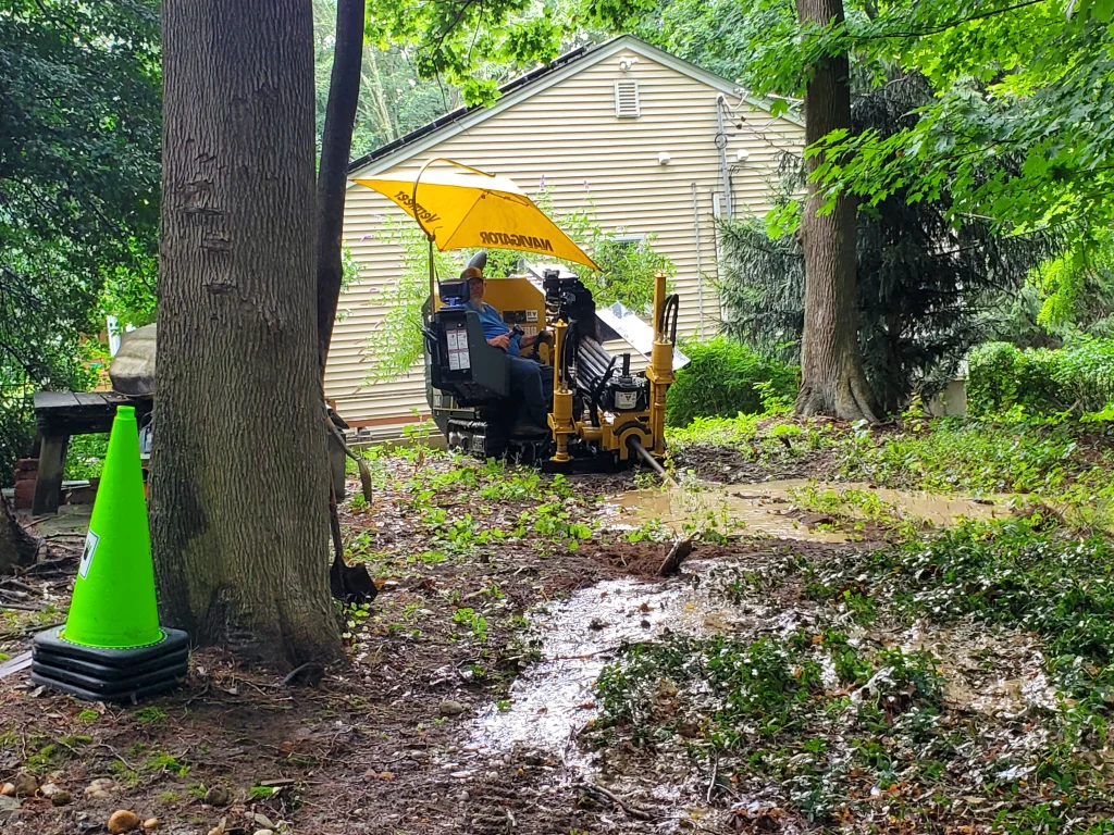 A worker operates a yellow Vermeer machine under a canopy in a muddy, wooded area near a house, with a bright green traffic cone visible in the foreground.