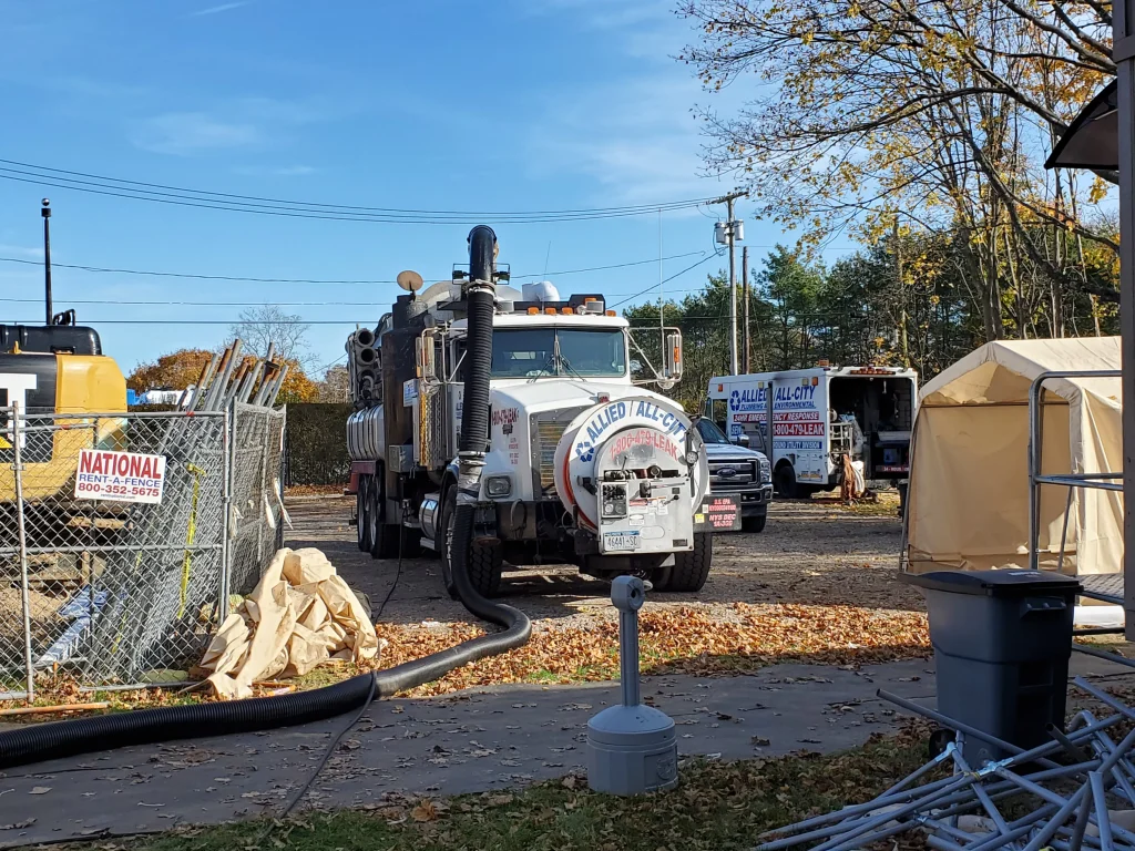 A large Allied Vacuum truck is parked at a construction site, with hoses connected to it. Nearby are construction materials, a fenced area, and trees with autumn leaves. Two Allied company vehicles are also visible in the background.