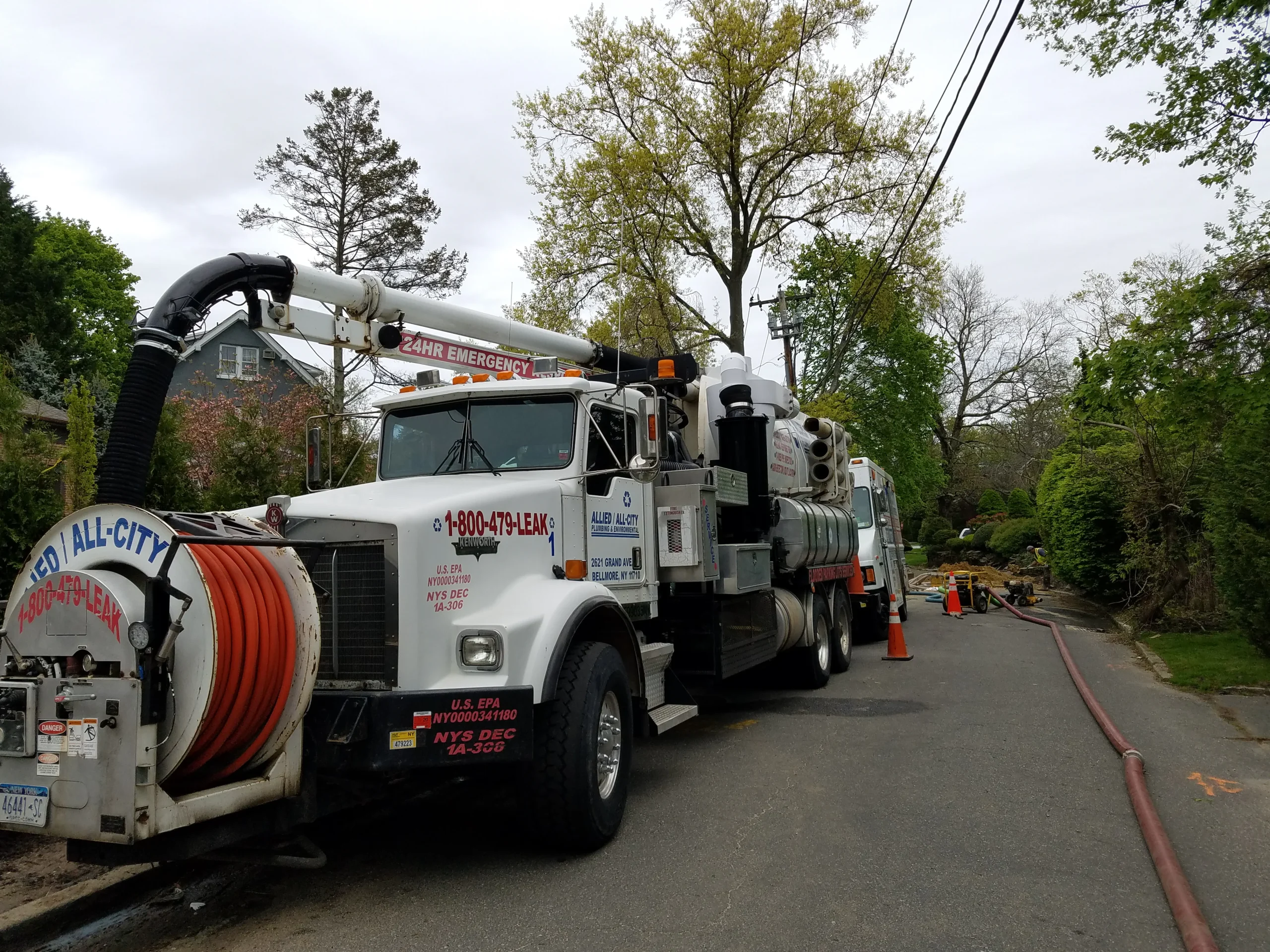 A large white utility truck with hoses and equipment is parked on a residential street. Orange cones and hoses stretch along the road, with workers and trees visible in the background.